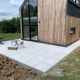 Nowoczesny dom z elewacją z naturalnego drewna i tarasem wyłożonym jasnymi płytami, na którym stoi stolik z krzesłami i grill. Widoczna świeżo przekopana ziemia wokół tarasu.