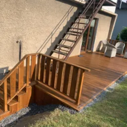 Nowoczesny taras z brązowych desek kompozytowych, z drewnianymi schodami prowadzącymi na trawnik i metalowymi schodami na piętro, wyposażony w szare rattanowe krzesła.