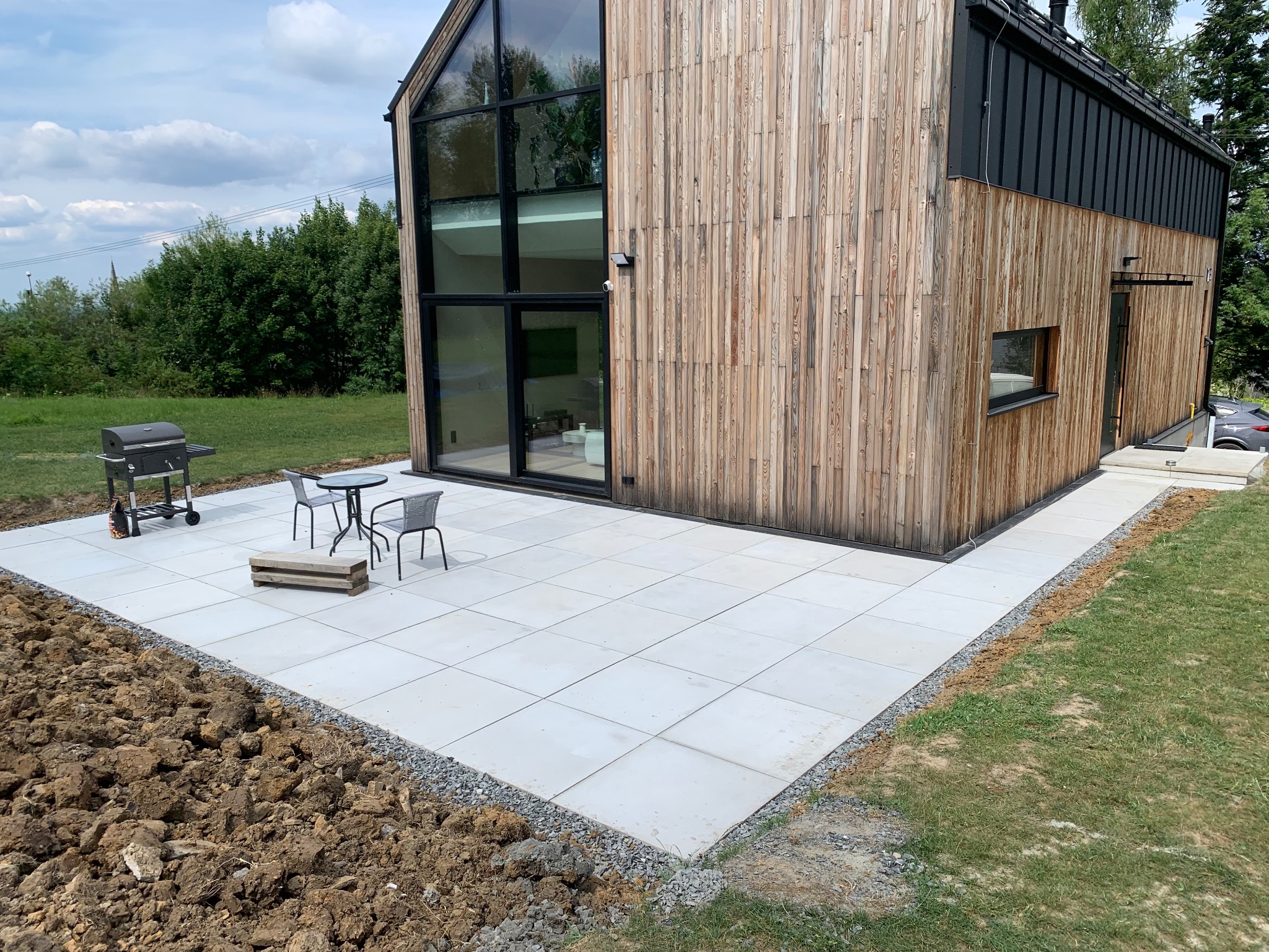 Nowoczesny dom z elewacją z naturalnego drewna i tarasem wyłożonym jasnymi płytami, na którym stoi stolik z krzesłami i grill. Widoczna świeżo przekopana ziemia wokół tarasu.