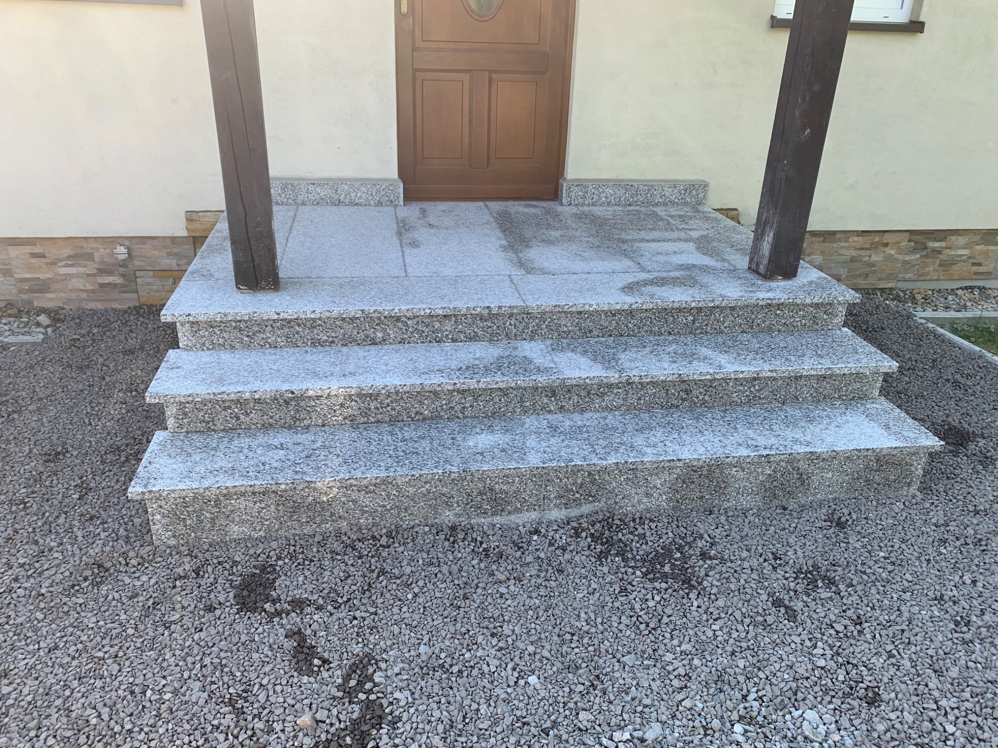 Granitowe schody wejściowe z trzema stopniami, prowadzące do drewnianych drzwi wejściowych, otoczone szarym żwirem i wsparte na ciemnych, drewnianych słupach.