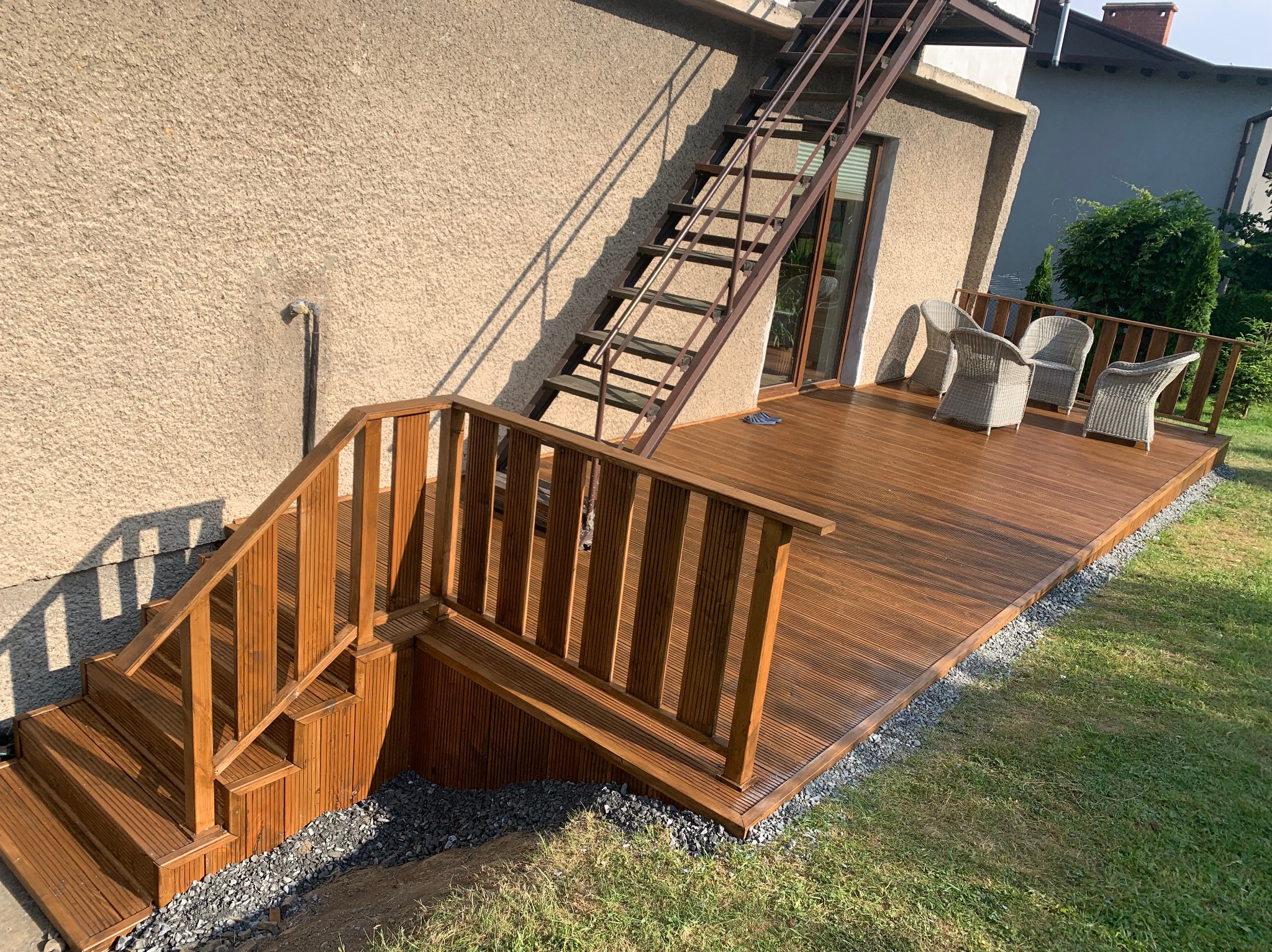 Nowoczesny taras z brązowych desek kompozytowych, z drewnianymi schodami prowadzącymi na trawnik i metalowymi schodami na piętro, wyposażony w szare rattanowe krzesła.