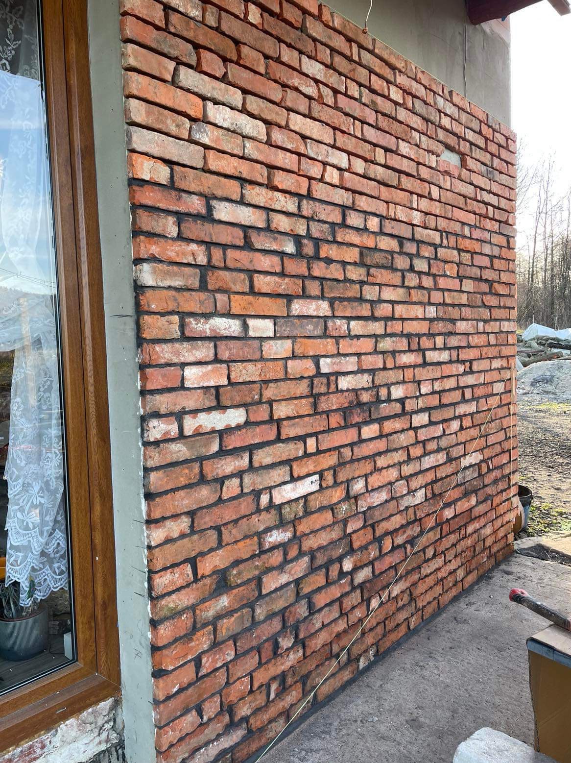 Ściana zewnętrzna budynku wykończona czerwoną cegłą klinkierową, widoczna obok okna z drewnianą ramą i firanką, z surowym betonowym wykończeniem narożnika.