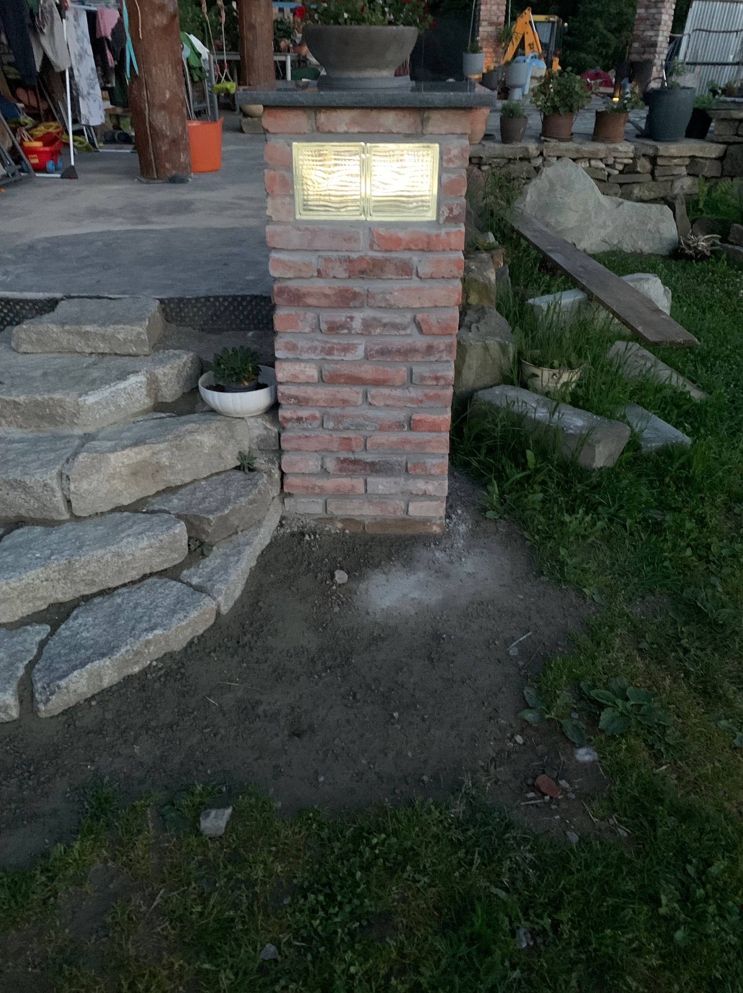 Kamienne schody prowadzące do tarasu z ceglanym słupkiem oświetleniowym, widoczny fragment trawnika i elementy ogrodu w tle.