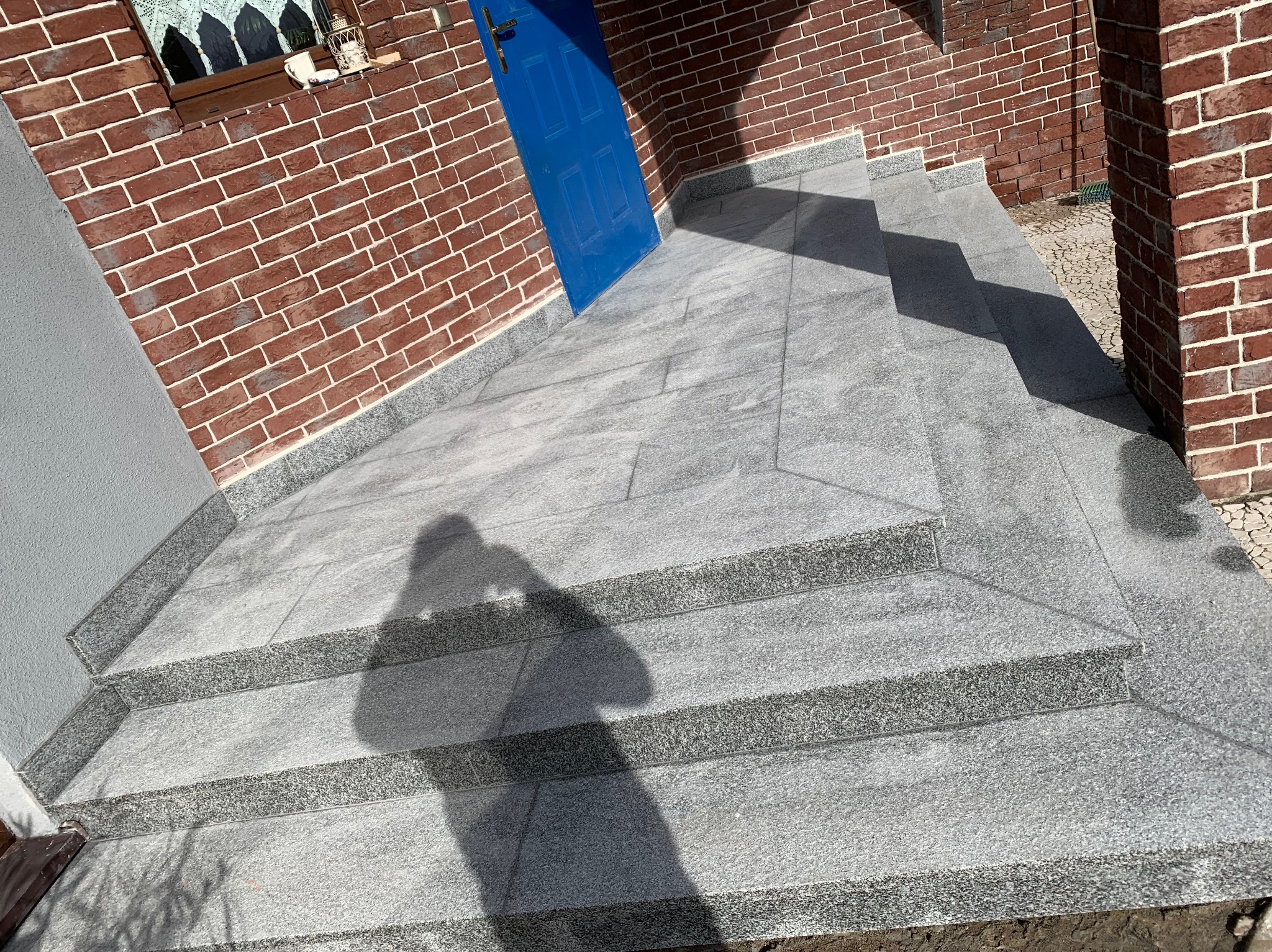 Zewnętrzne schody wejściowe wyłożone szarymi płytami kamiennymi, z widocznym cieniem osoby robiącej zdjęcie, prowadzące do niebieskich drzwi wejściowych w budynku z cegły.