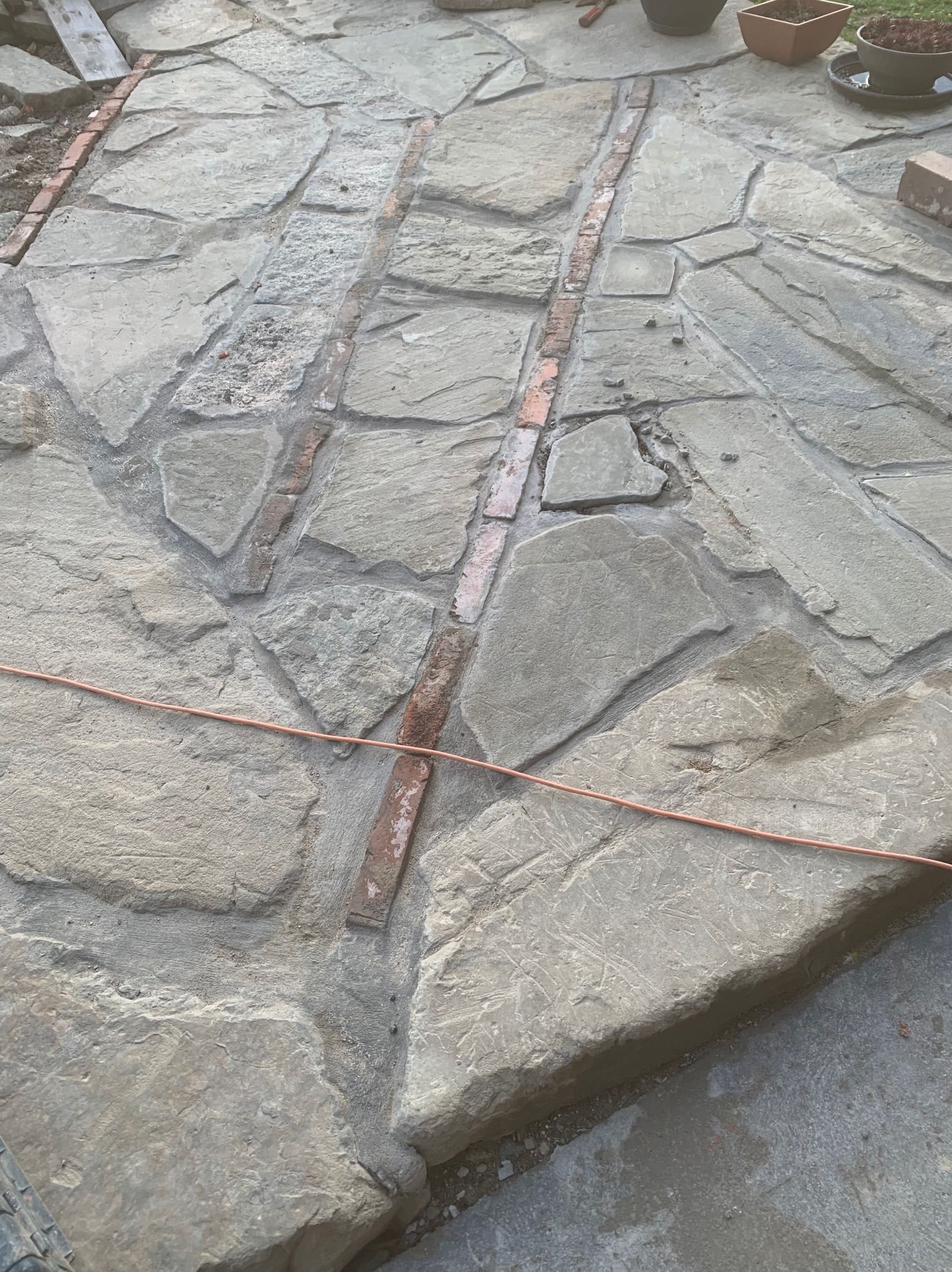 Nierówna kamienna powierzchnia w trakcie budowy, z widocznymi fragmentami cegieł oddzielającymi nieregularne płyty, na tle widoczne doniczki z roślinami.