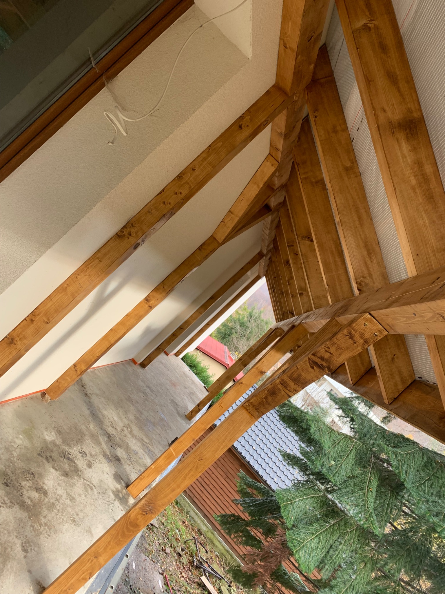 Wnętrze w trakcie budowy z widoczną drewnianą konstrukcją dachu i ścian, surowy beton na podłodze, widok na zewnątrz na drzewa i budynki.