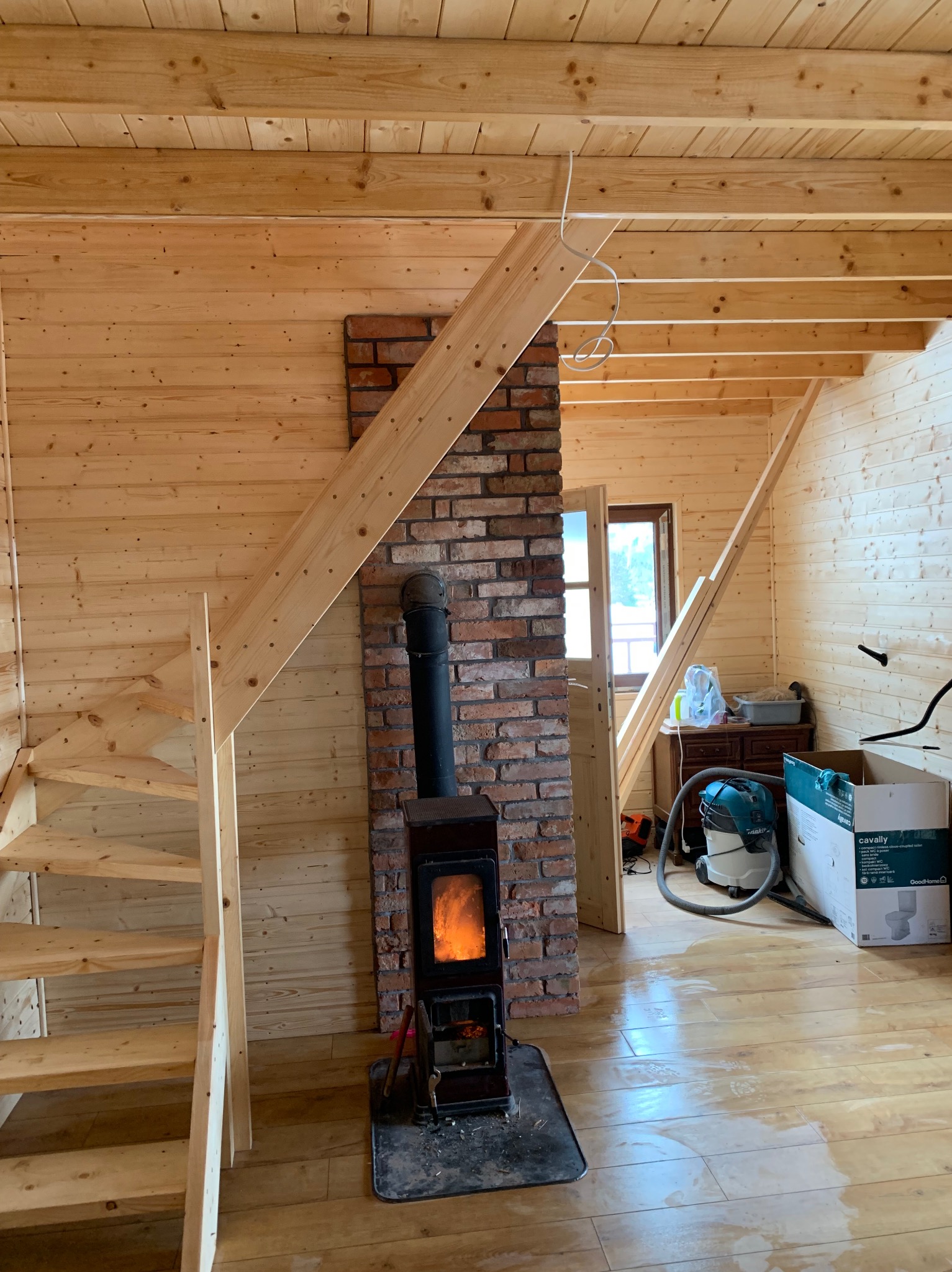 Wnętrze drewnianego domu w trakcie wykańczania z piecem kominkowym na tle ceglanej ściany, widoczne surowe drewniane schody i podłoga z połyskiem, w tle narzędzia i kartony.