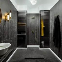 Elegancka łazienka w ciemnej tonacji z mozaikową podłogą, złotymi akcentami w postaci armatury i lustra, oraz kabiną prysznicową z czarnymi ramami.