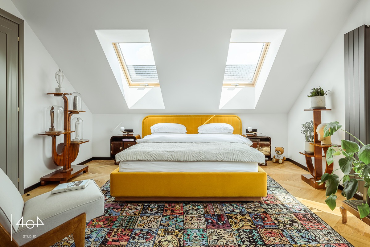 Jasna sypialnia na poddaszu z żółtym łóżkiem, wzorzystym dywanem, drewnianą podłogą w jodełkę i dwoma oknami dachowymi. Uwagę przykuwają oryginalne, drewniane półki na dekoracje i rośliny.