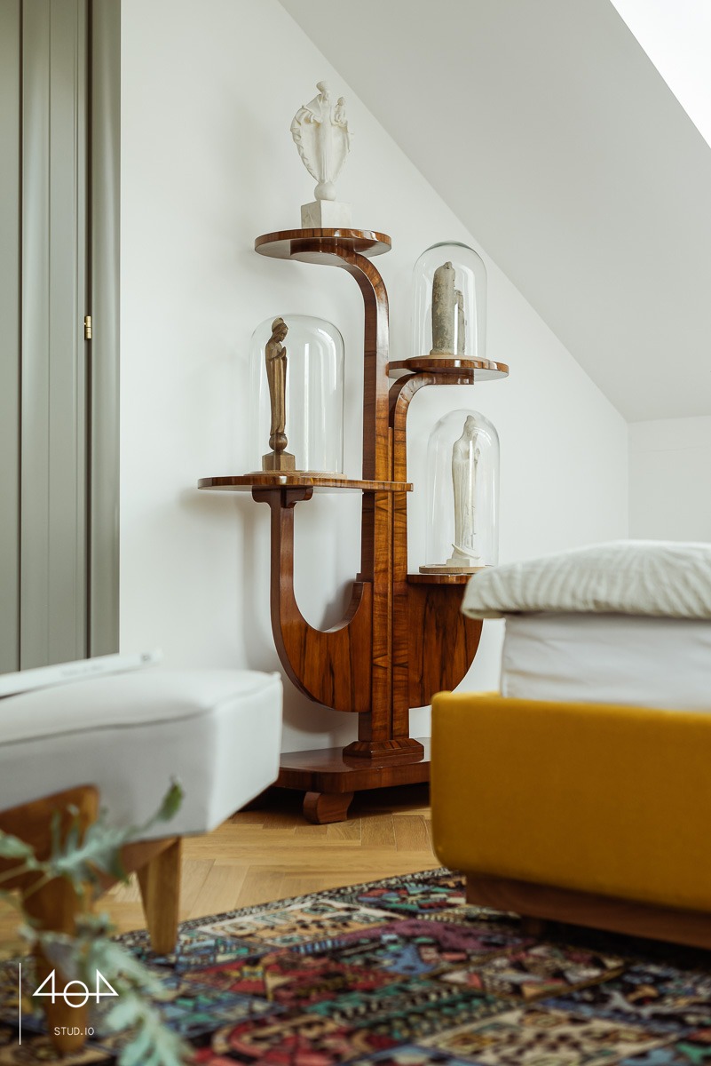 Wysoki, artystyczny regał z drewna o ciemnym usłojeniu, z półkami eksponującymi rzeźby pod szklanymi kloszami, ustawiony w jasnym pokoju na tle białej ściany i dywanu w orientalne wzory.