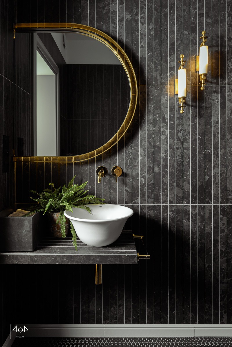 Elegancka łazienka w ciemnej tonacji: biała umywalka typu miska, złote detale armatury i ramy lustra, marmurowe płytki na ścianie, akcent zieleni w postaci paprotki.