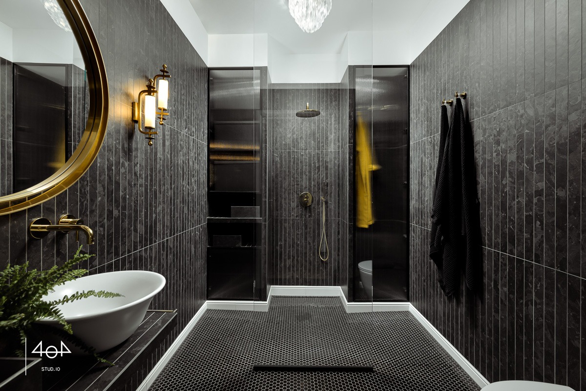 Elegancka łazienka w ciemnej tonacji z mozaikową podłogą, złotymi akcentami w postaci armatury i lustra, oraz kabiną prysznicową z czarnymi ramami.