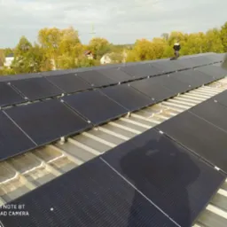 Instalacja paneli fotowoltaicznych na metalowym dachu z widocznym pracownikiem w odzieży roboczej i drzewami w tle.