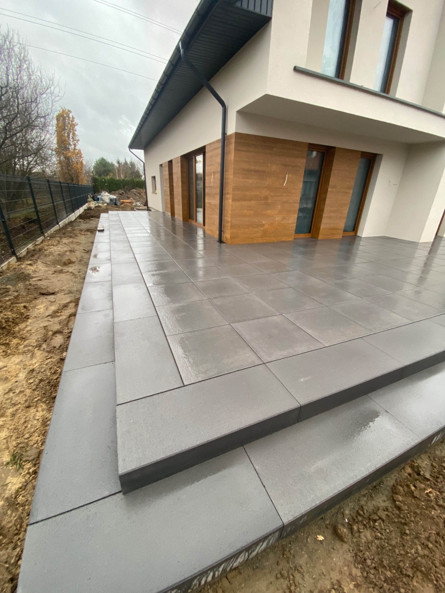 Nowoczesny taras z szarej kostki brukowej prowadzący do domu z drewnianą elewacją, widoczne dwa stopnie i fragment ogrodu z metalowym ogrodzeniem.