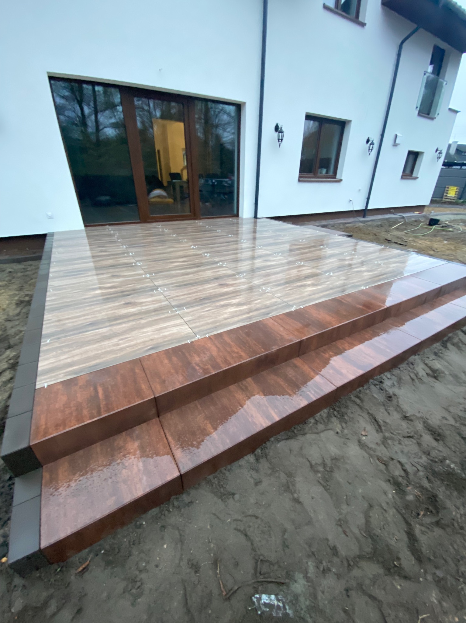 Wyłożony płytkami taras z dwoma schodkami przed nowoczesnym domem, widoczne krzyżyki dystansowe między płytkami, mokra powierzchnia.