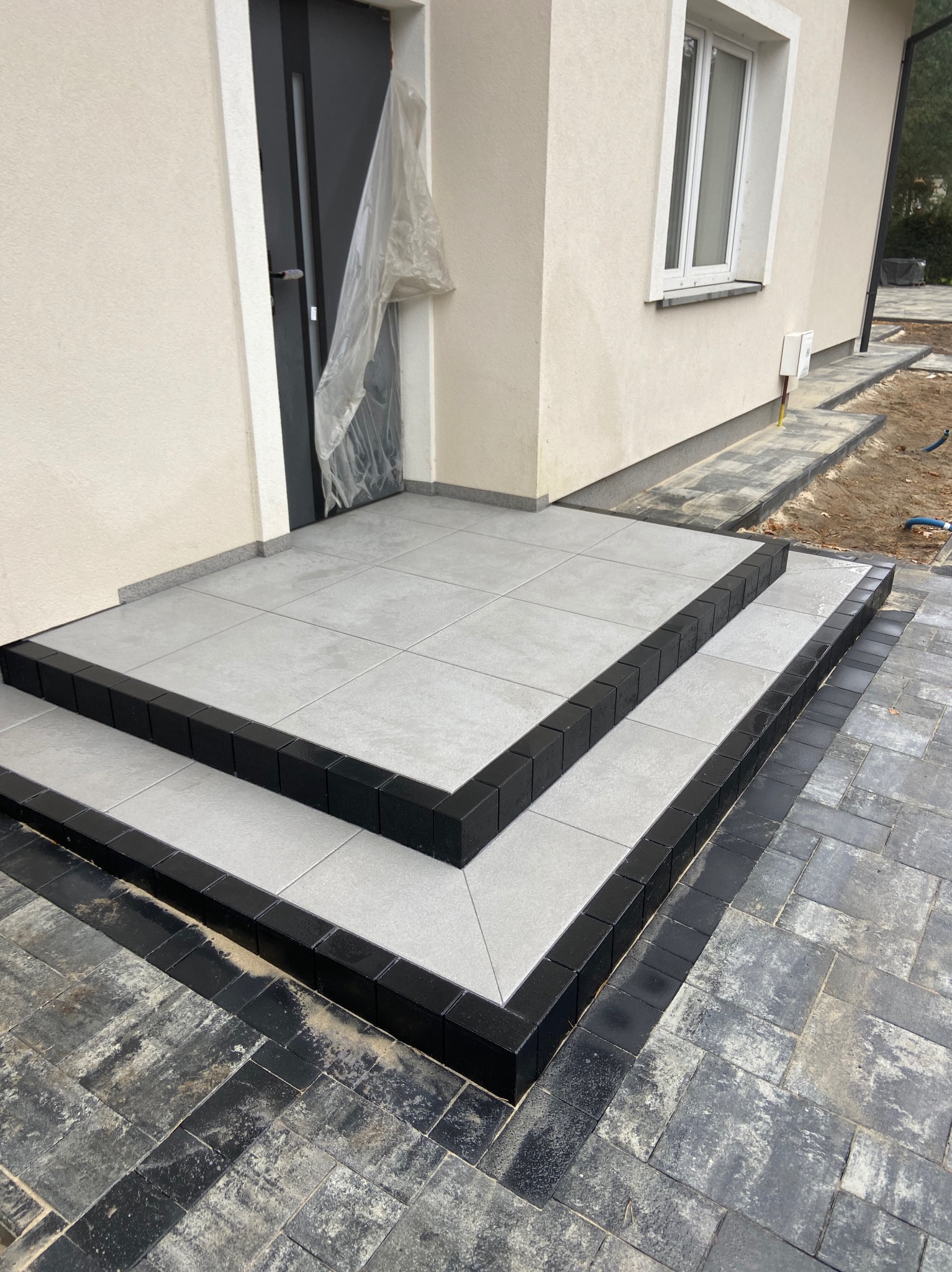 Schody wejściowe wyłożone jasnoszarą płytką ceramiczną z czarnym obramowaniem, prowadzące do drzwi zabezpieczonych folią, obok ułożona kostka brukowa w odcieniach szarości i czerni.