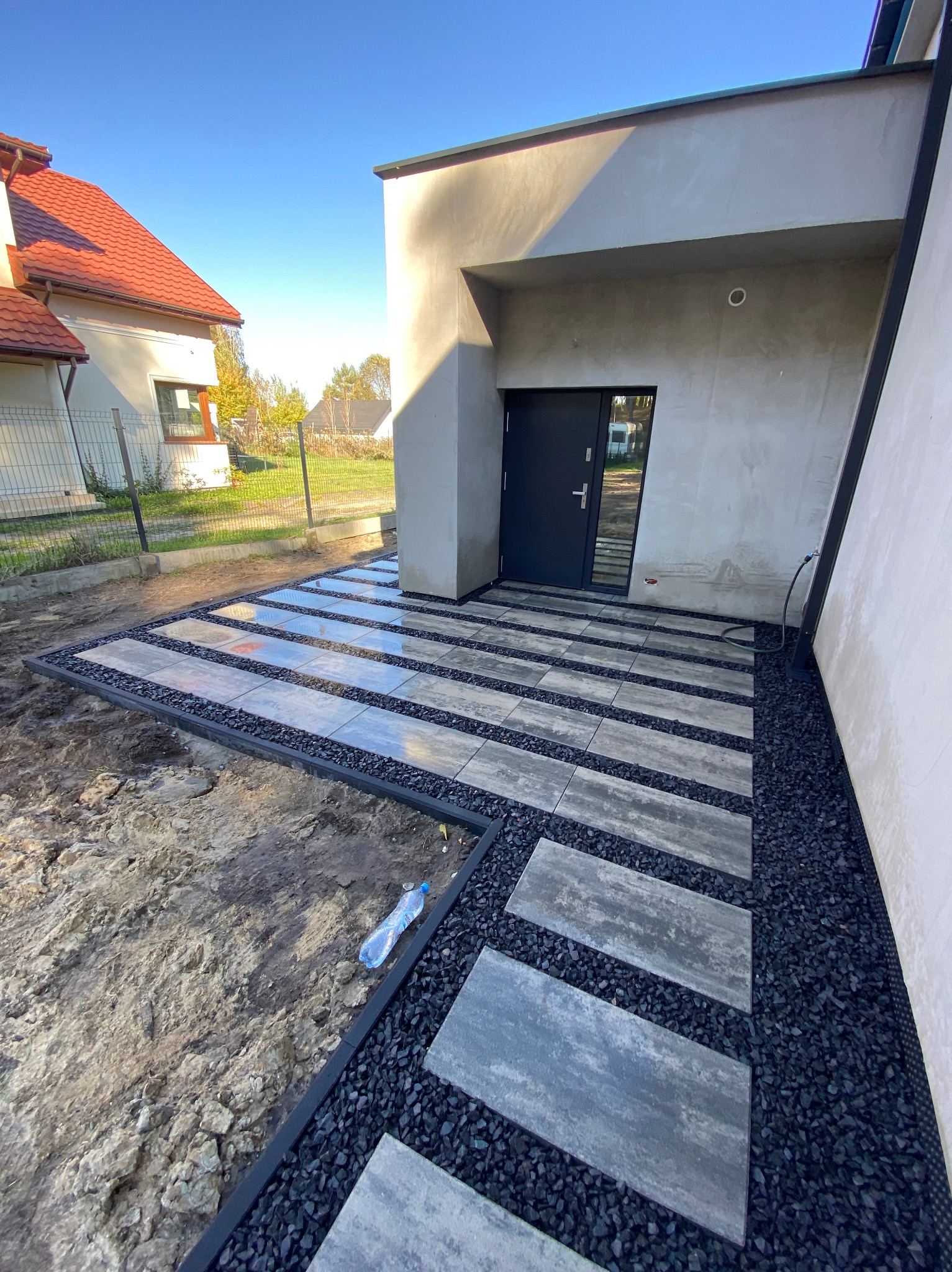 Nowoczesna ścieżka z szarych płyt betonowych oddzielonych czarnym grysem, prowadząca do drzwi wejściowych domu o minimalistycznej architekturze. W tle widoczny dom z czerwoną dachówką.