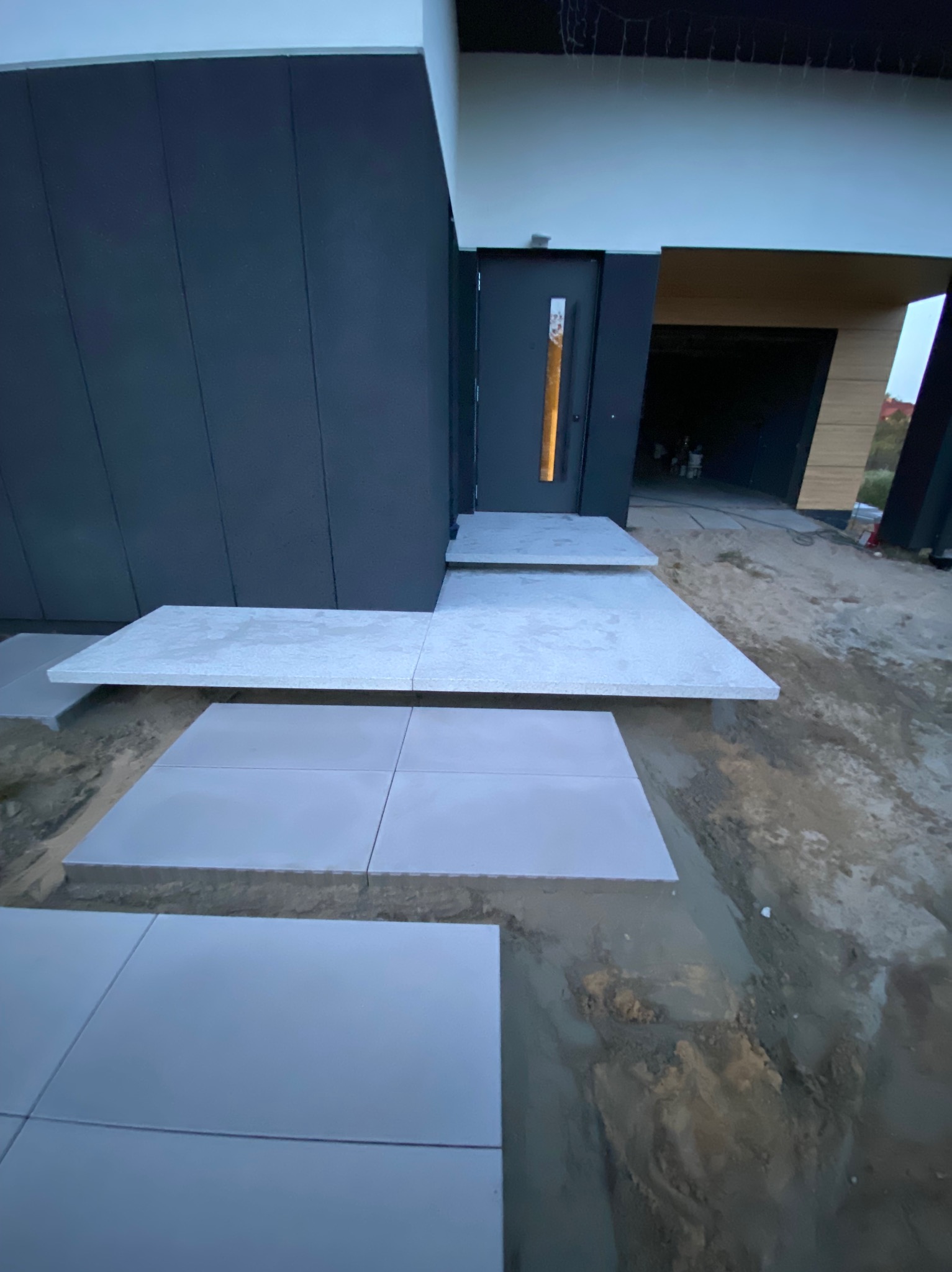 Nowoczesne wejście do domu z szarych płyt chodnikowych, ułożonych w formie schodów i ścieżki, prowadzące do ciemnych drzwi wejściowych z podświetlanym numerem.