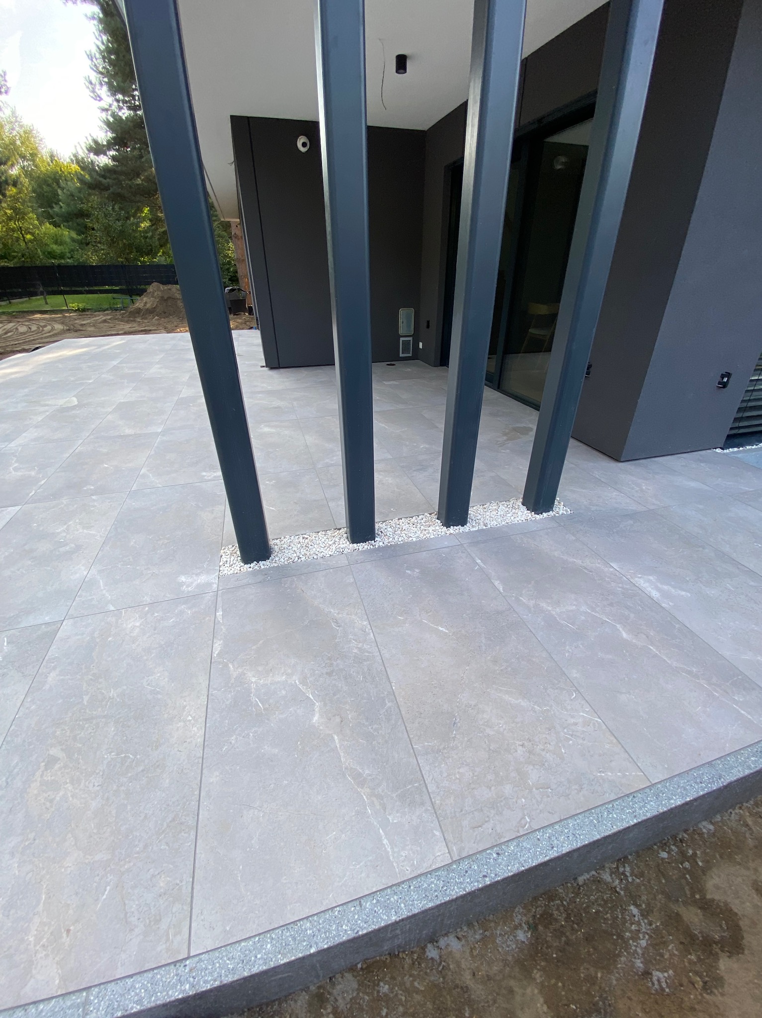 Jasnoszara, marmurkowa posadzka tarasowa z czterema pionowymi, grafitowymi słupami, otoczonymi białym grysem, na tle nowoczesnego domu w ciemnych kolorach.