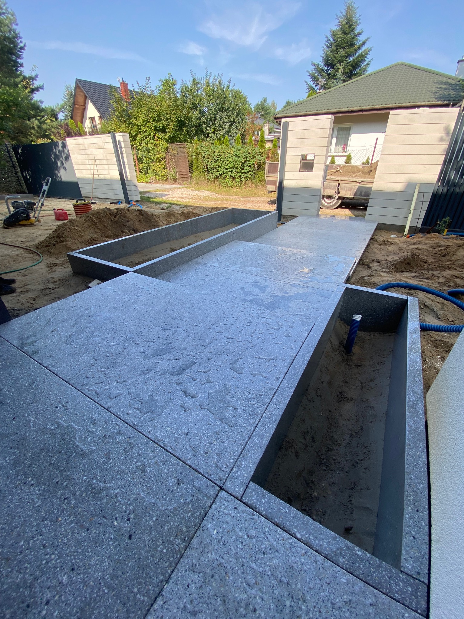 Świeżo ułożona, mokra, szara nawierzchnia z płyt granitowych w ogrodzie, obramowana obrzeżami, z widocznymi wykopanymi rabatami i sprzętem budowlanym w tle.