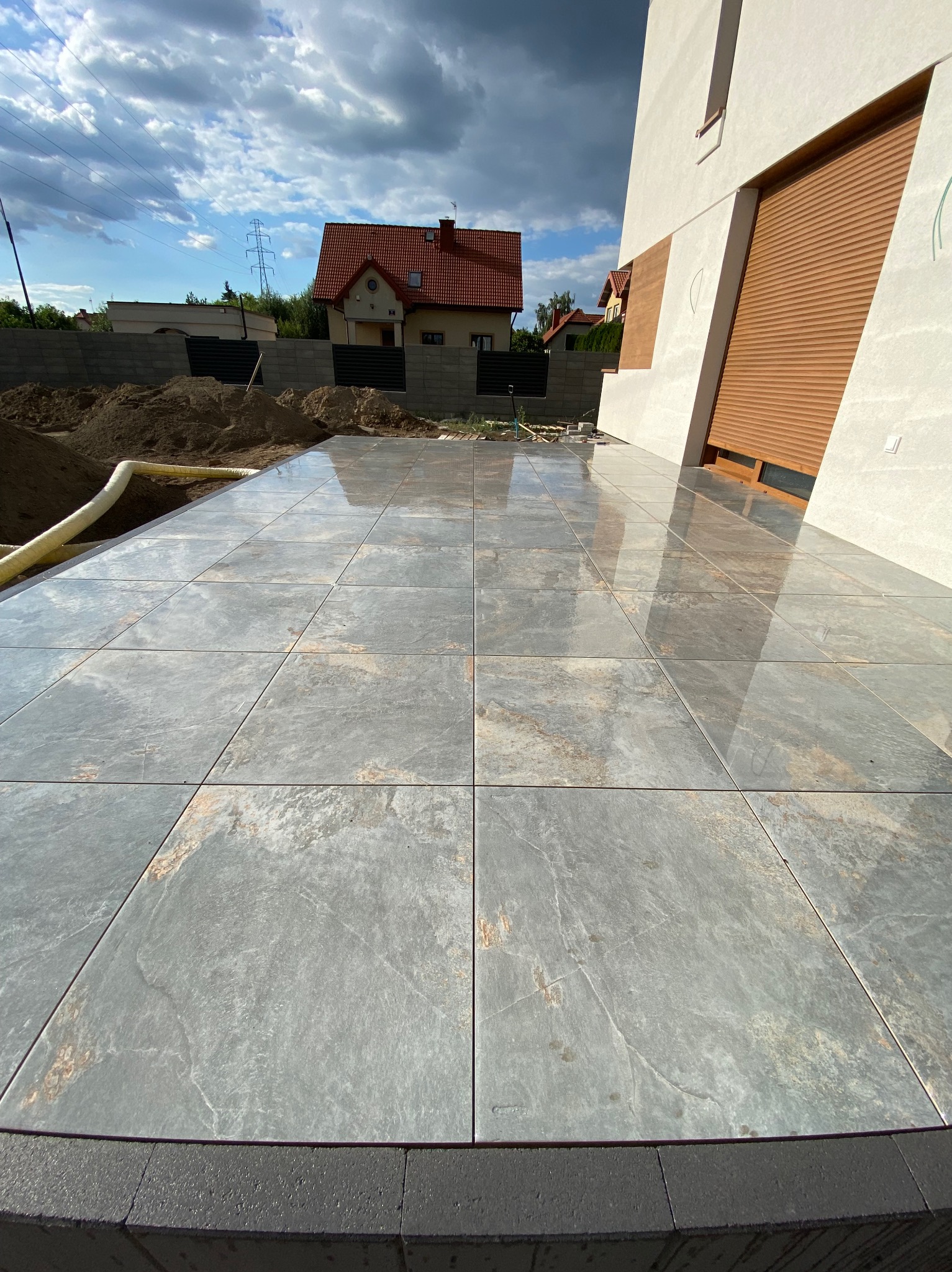Ułożona, mokra, szara kostka na tarasie domu jednorodzinnego, widok na dom z roletami i stertę ziemi w tle.