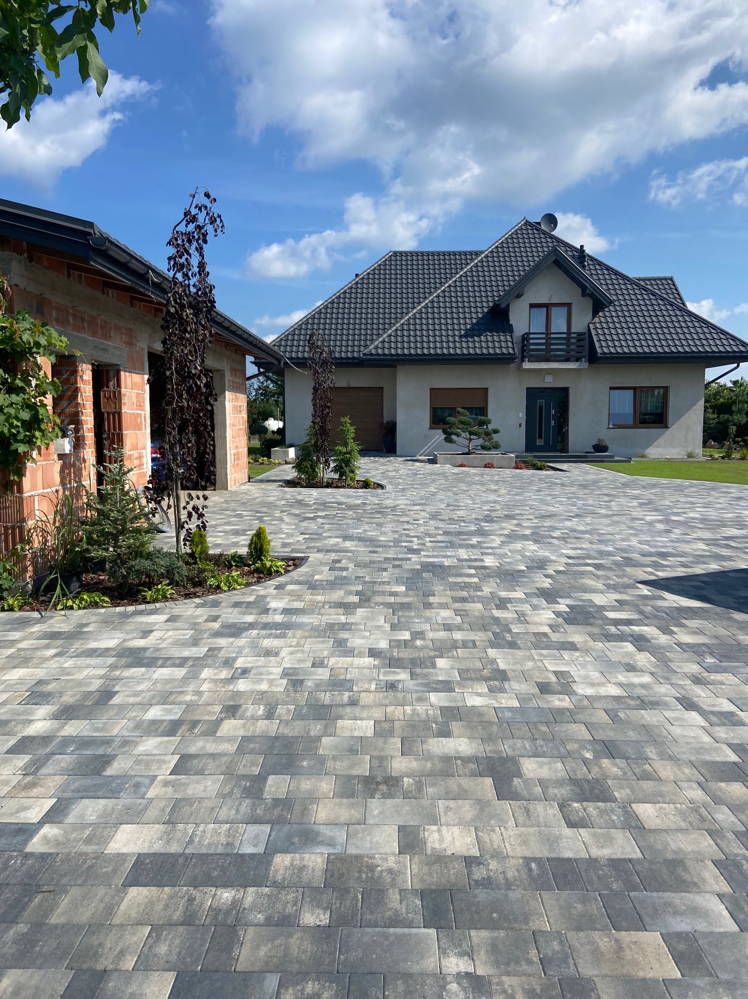 Szeroki podjazd z szarej kostki brukowej prowadzący do nowoczesnego domu z grafitowym dachem i fragmentu budynku w trakcie budowy z czerwonej cegły, otoczonego zielenią.