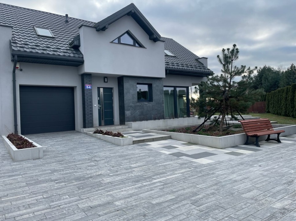 Nowoczesny dom jednorodzinny z podjazdem wyłożonym szarą kostką w podłużnym wzorze, rabaty kwiatowe w betonowych obrzeżach i drewniana ławka obok niedużego drzewka.
