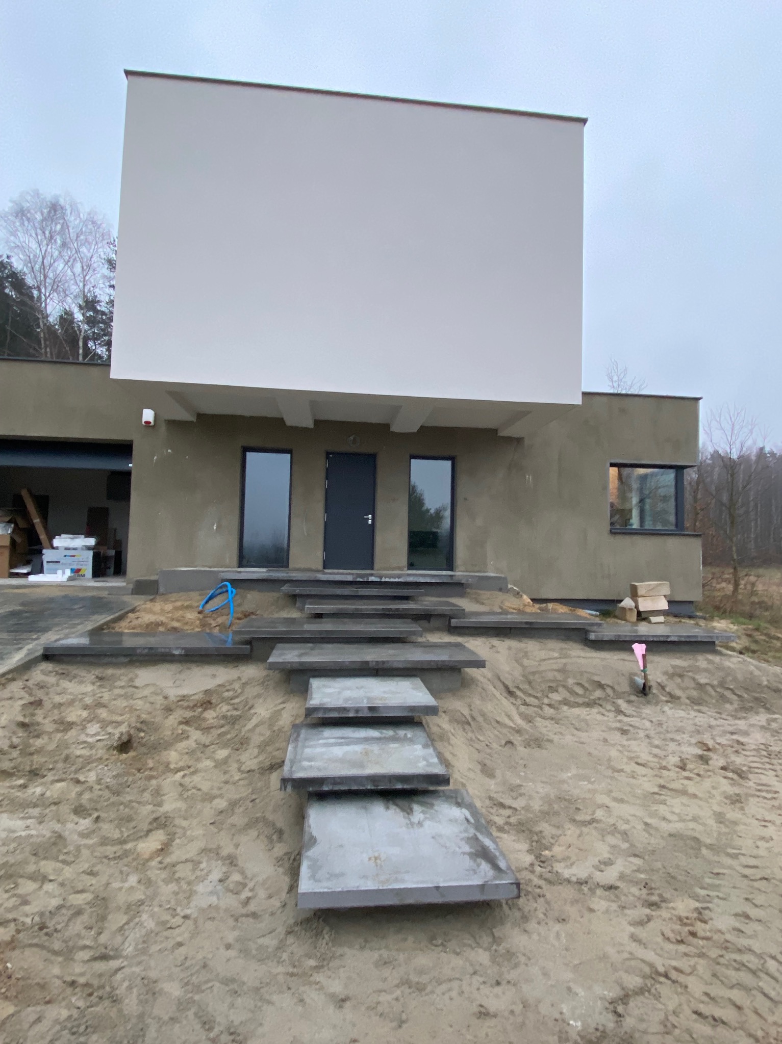 Nowoczesny dom z szarymi, betonowymi stopniami prowadzącymi do wejścia, teren wokół w trakcie prac ziemnych, widoczna niebieska rura.