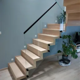 Nowoczesne schody z jasnego drewna z przezroczystą szklaną balustradą mocowaną czarnymi elementami, na tle szarej ściany i drewnianej podłogi, w pobliżu rośliny doniczkowej i legowiska dla zwierząt.