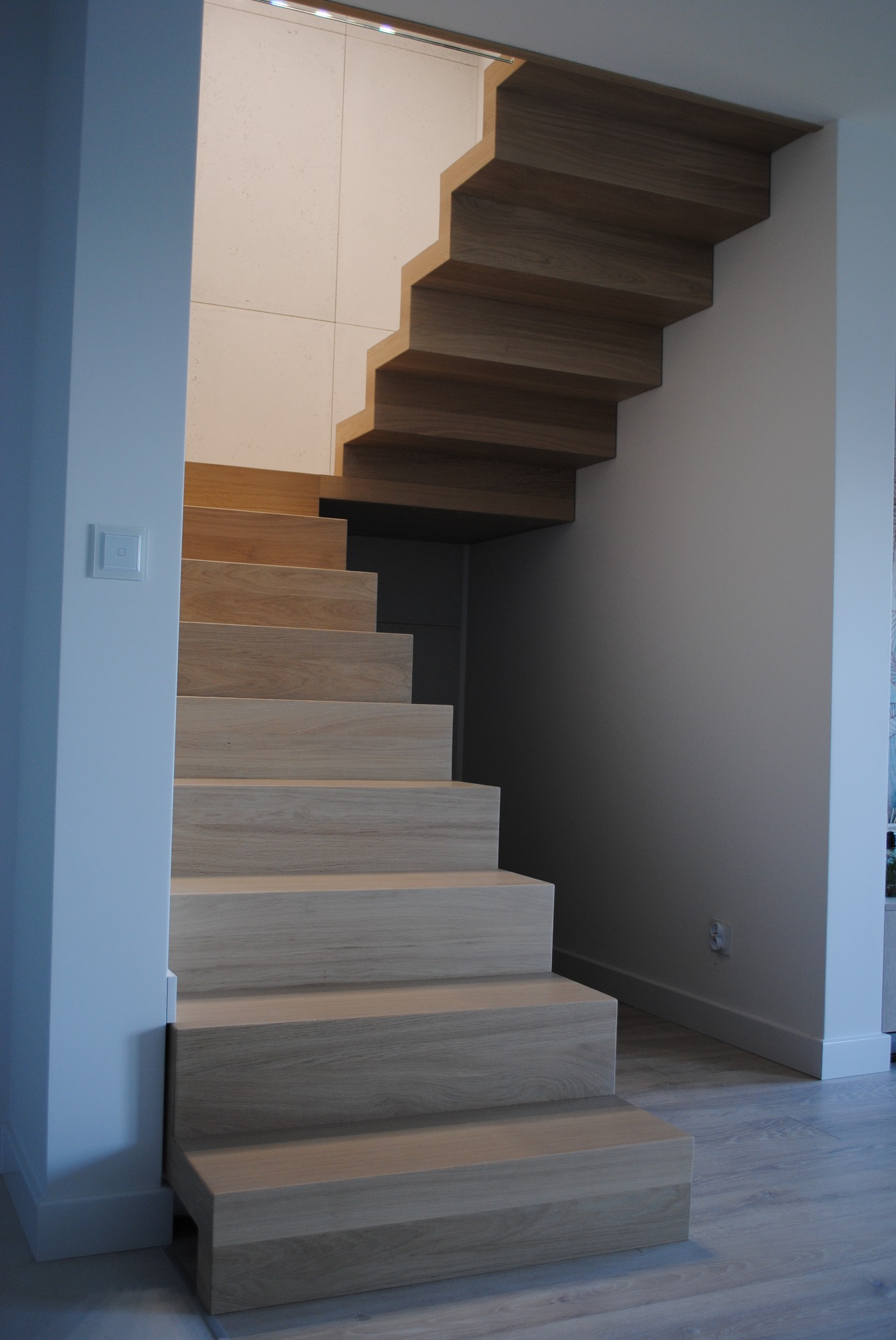 Minimalistyczne schody z jasnego drewna, bez poręczy, z widocznymi usłojeniami, zamontowane przy białej ścianie z włącznikiem światła.