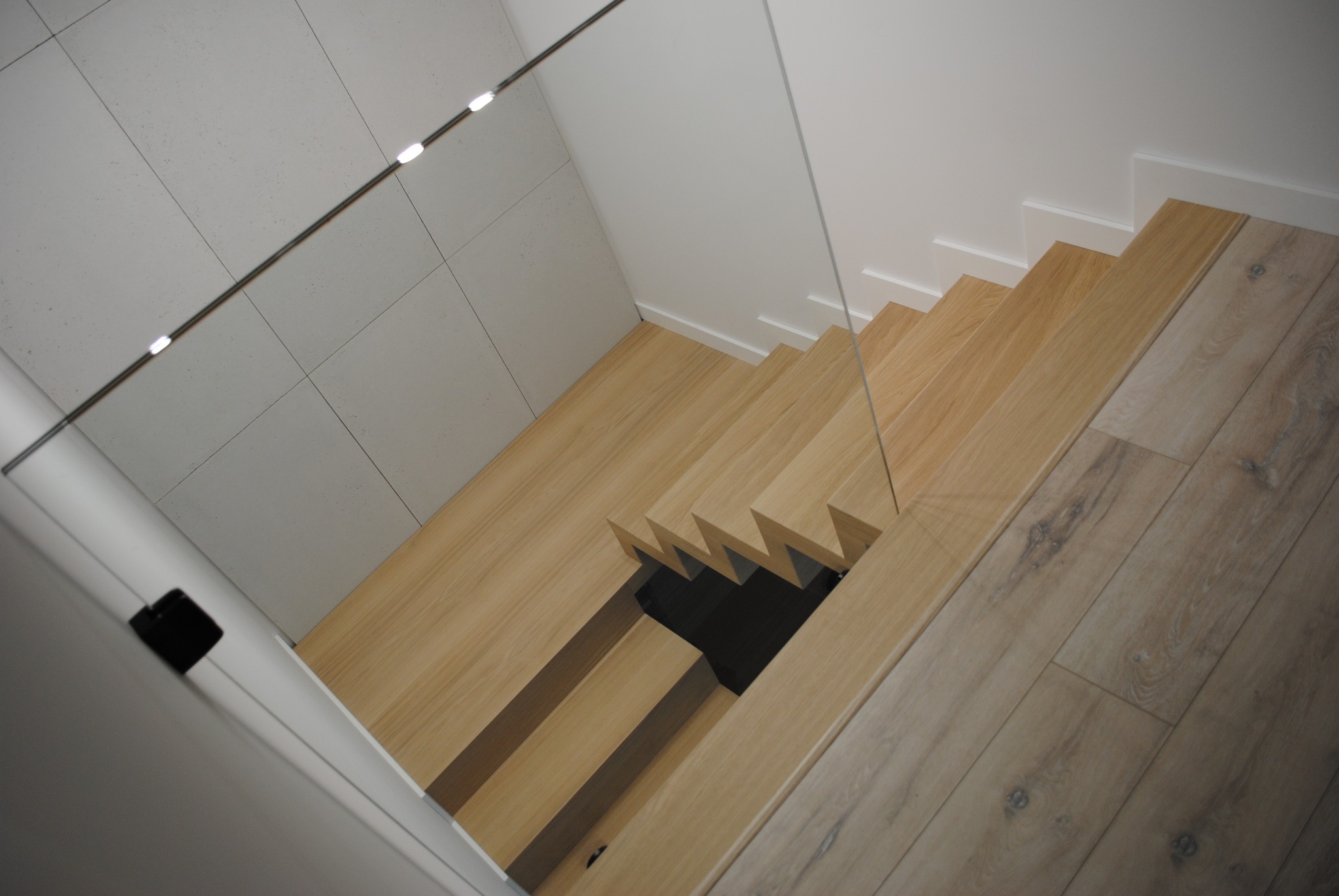 Nowoczesne schody drewniane z jasnego drewna, zabezpieczone szklaną balustradą, widok z góry, minimalistyczny design z białymi ścianami i podświetleniem.