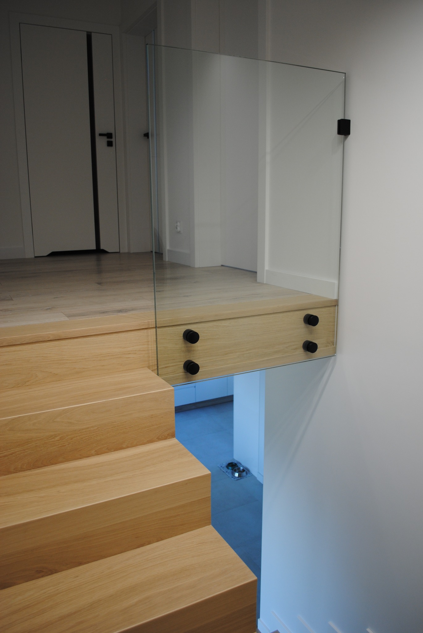 Nowoczesne schody z jasnego drewna z szklaną balustradą mocowaną na czarnych uchwytach, widok z góry na dół.