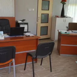 Biuro z dwoma biurkami z laptopami, drukarką i krzesłami na kafelkowej podłodze, w tle szafka z kwiatami w wazonie.