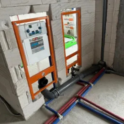 Widok na surową ścianę z betonu komórkowego z dwoma wnękami z pomarańczowymi ramami montażowymi pod zabudowę WC, widoczne szare rury kanalizacyjne i czerwono-niebieskie rury instalacji wodnej...