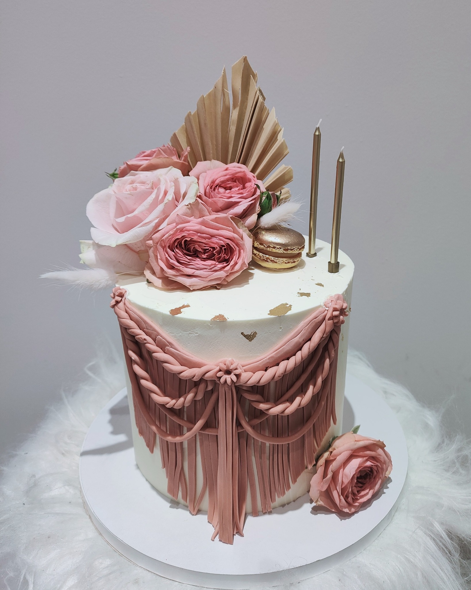 Tort zdobiony różowymi różami, złotym makaronikiem i dekoracją z masy imitującą drapowaną tkaninę w odcieniu pudrowego różu, ustawiony na białym, puszystym podłożu.