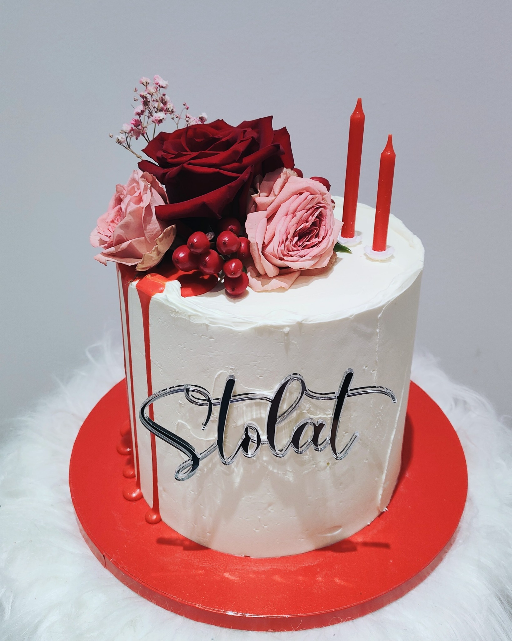 Tort urodzinowy z białym kremem, czerwonymi świeczkami, dekoracją z czerwonych i różowych róż oraz napisem 'Stolat' w srebrnym kolorze, całość umieszczona na czerwonym talerzu na białym puszystym...