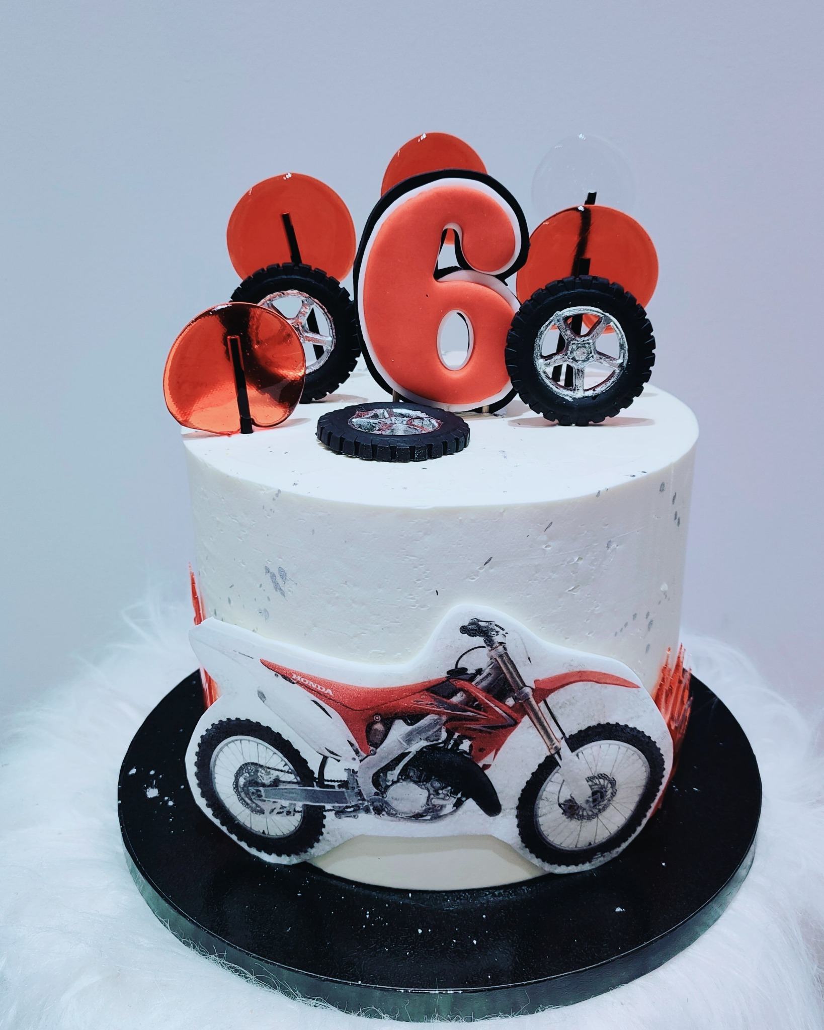 Tort urodzinowy z motywem motocykla Honda, udekorowany cyfrą '6', kołami i czerwonymi lizakami na białym tle.