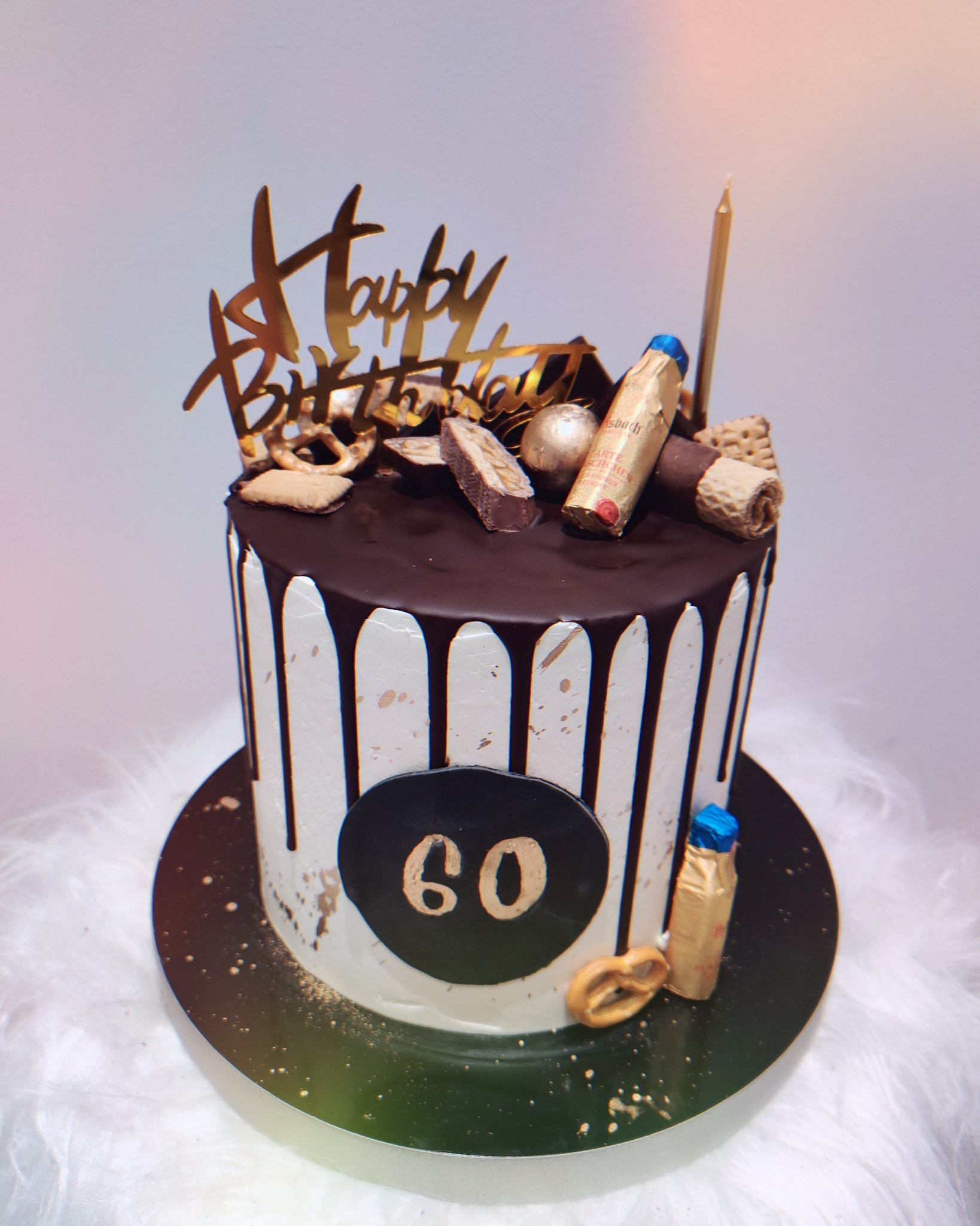 Tort urodzinowy z polewą czekoladową, dekoracjami z batoników, precli i złotym napisem 'Happy Birthday', świeczka na torcie, liczba '60' na froncie.
