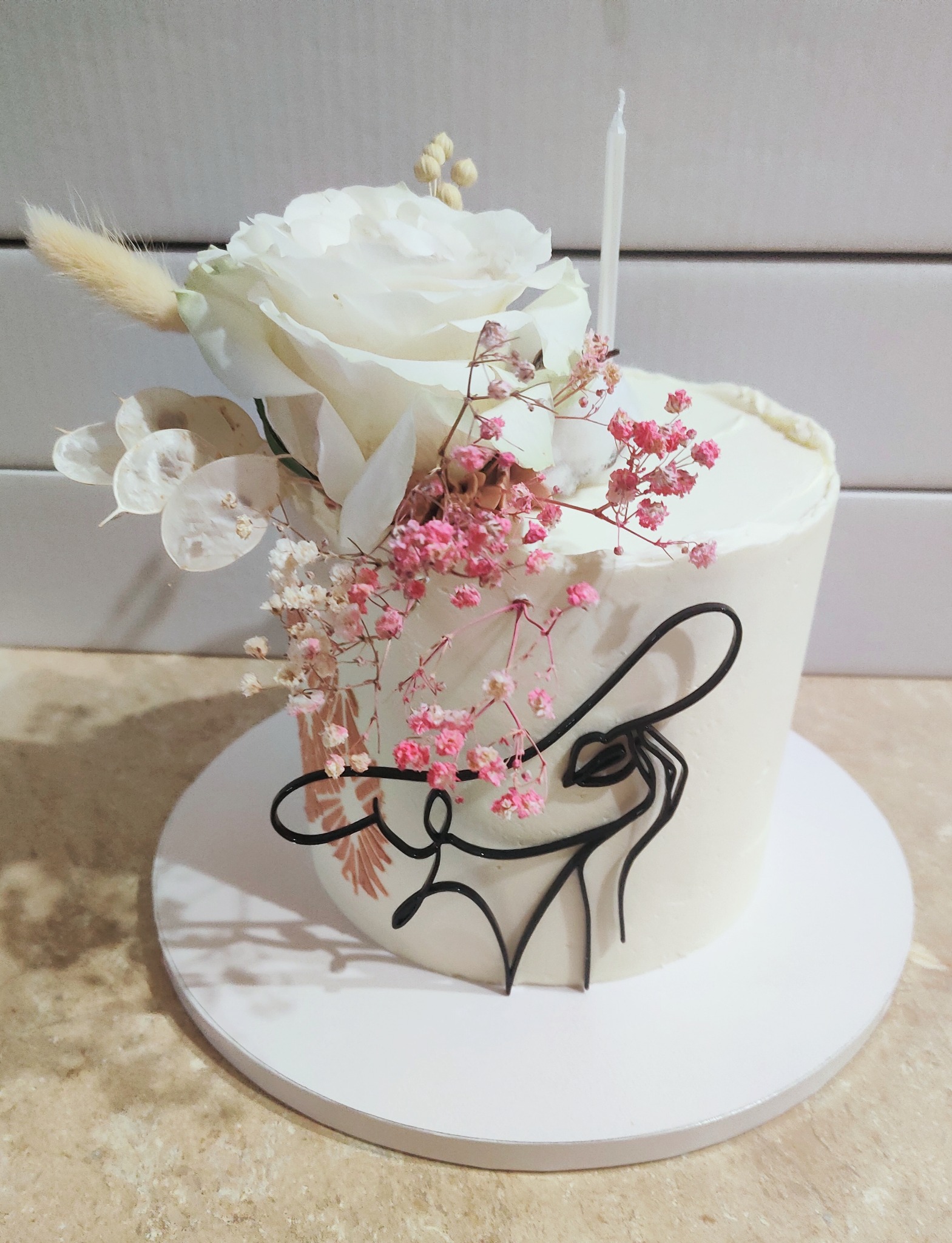 Tort z białym kremem, ozdobiony białą różą, drobnymi różowymi kwiatami i czarnym konturem twarzy kobiety w kapeluszu.