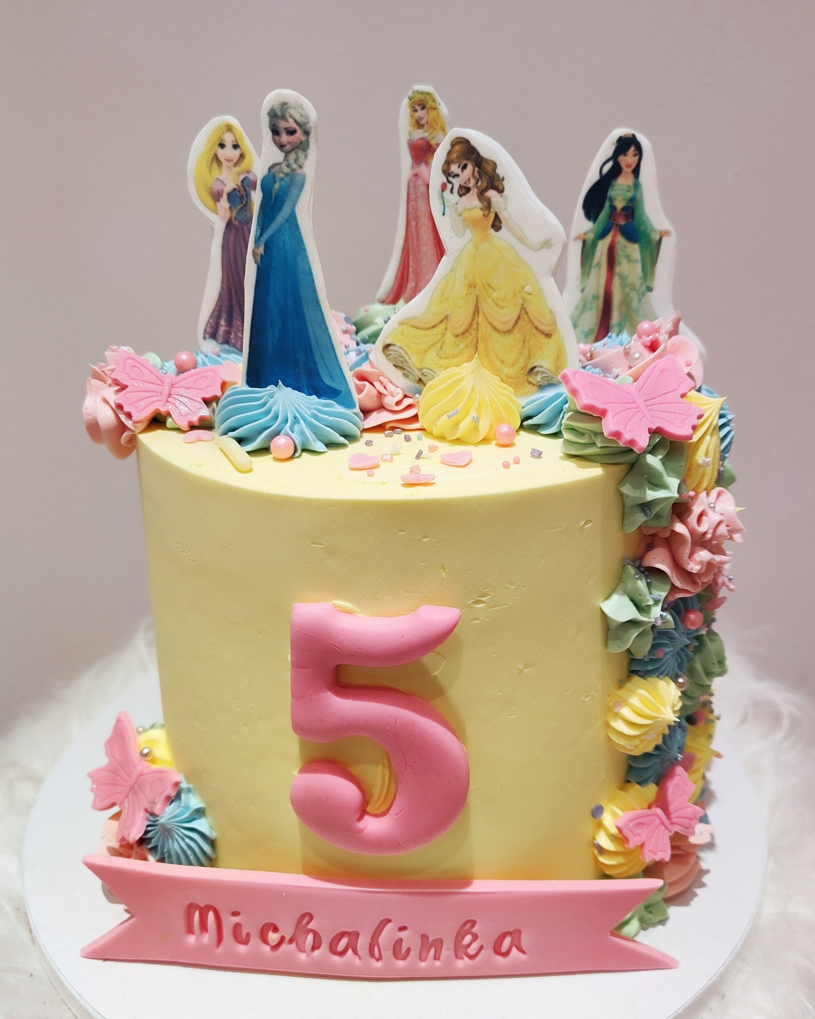 Tort urodzinowy w pastelowych kolorach z dekoracjami z lukru w kształcie kwiatów, motyli i figurek księżniczek Disneya, z różową cyfrą '5' i napisem 'Michalinka'.