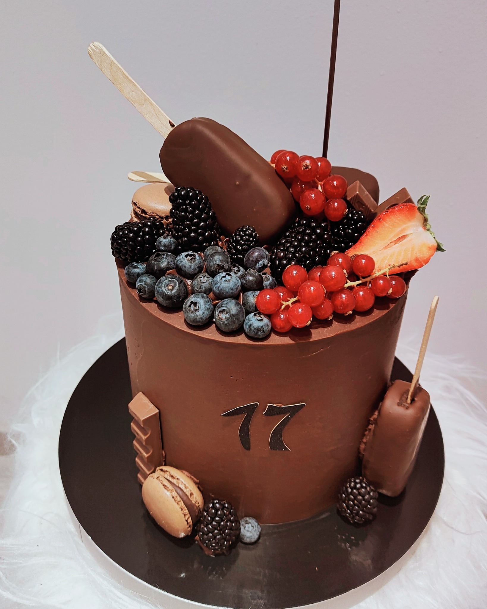 Czekoladowy tort urodzinowy z numerem 77, udekorowany świeżymi jagodami, borówkami, porzeczkami, truskawką, makaronikami i czekoladowymi lodami na patyku.