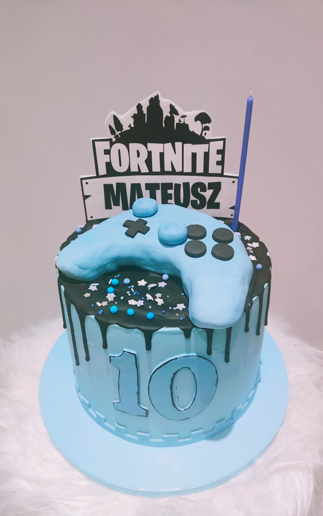 Tort urodzinowy w odcieniach błękitu i czerni, dekorowany motywem z gry Fortnite i kontrolerem, z napisem '10' na boku i świeczką.