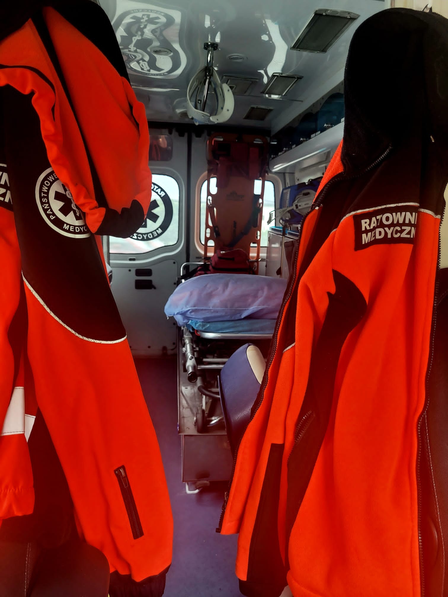 Wnętrze ambulansu z widocznymi wiszącymi kurtkami ratowników medycznych, noszami i sprzętem medycznym.