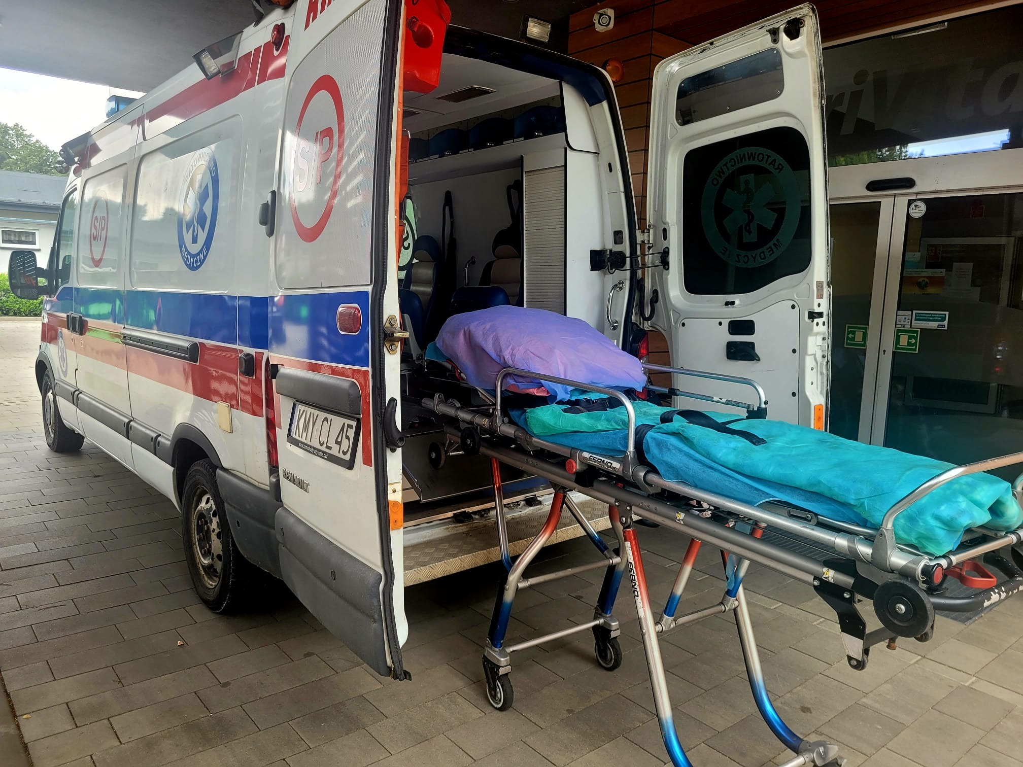 Otwarty ambulans z wysuniętymi noszami, przykrytymi niebieskim i fioletowym materiałem, zaparkowany przed budynkiem z automatycznymi drzwiami.