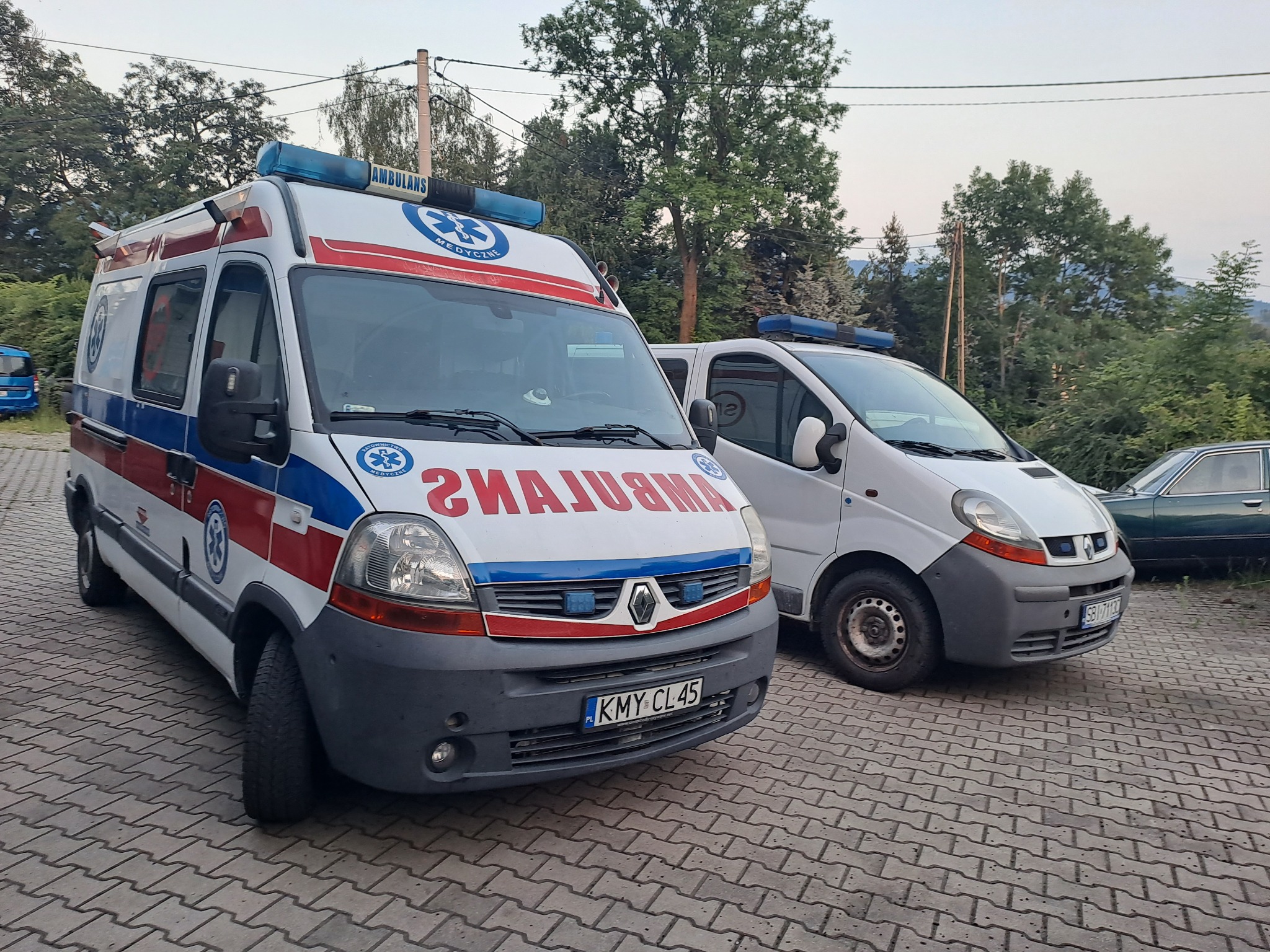 Dwa ambulanse zaparkowane na brukowanym placu, pierwszy z widocznym numerem rejestracyjnym KMY CL45 i oznaczeniami medycznymi, drugi częściowo zasłonięty, w tle drzewa i fragment samochodu osobowego.