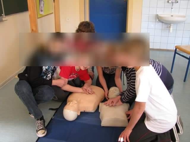 Grupa osób ćwiczy resuscytację krążeniowo-oddechową na manekinach na niebieskiej macie w jasnym pomieszczeniu z umywalką w tle.