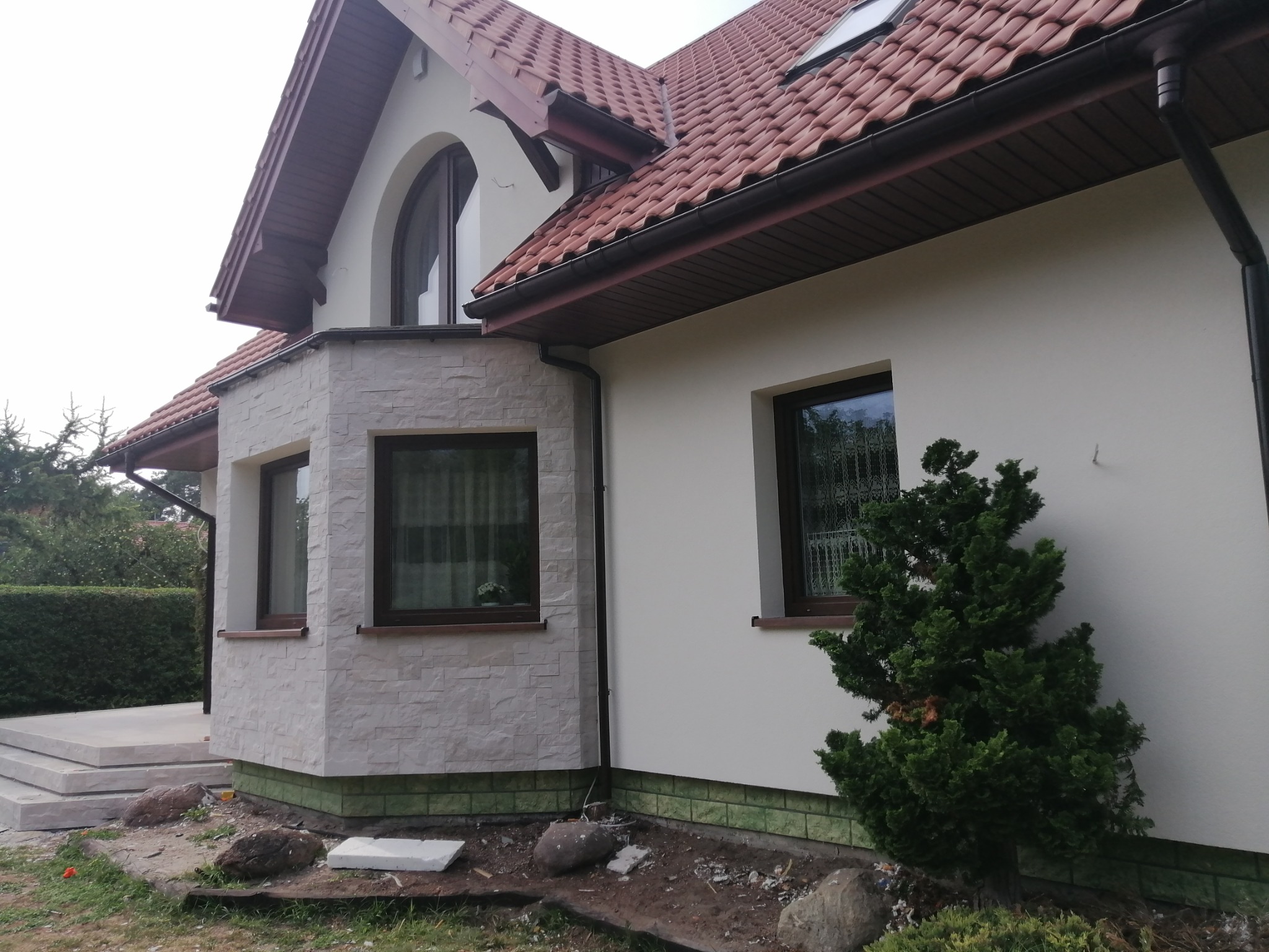 Jasna elewacja domu jednorodzinnego z fragmentem okładziny kamiennej, brązowe okna i dachówka ceramiczna, widoczne prace wykończeniowe wokół budynku.