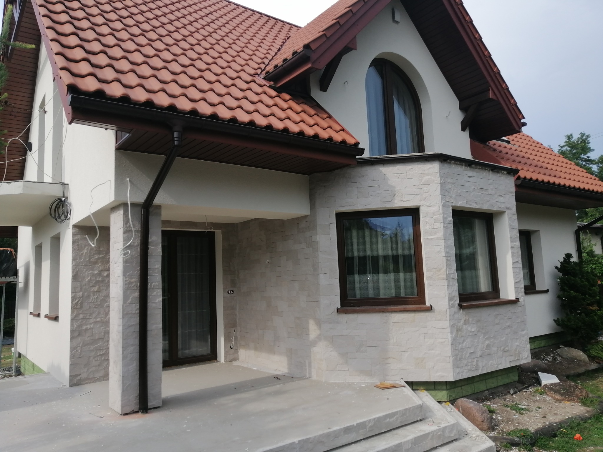 Elewacja domu jednorodzinnego z wykończeniem z jasnego kamienia dekoracyjnego, brązowa dachówka, widoczne elementy orynnowania i okna z brązowymi ramami.