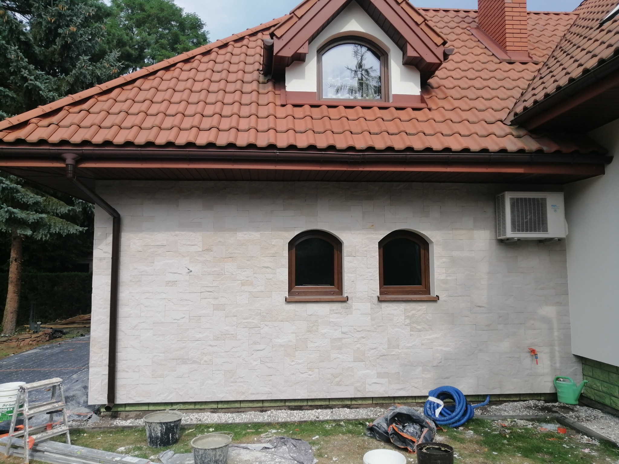 Wykończona elewacja domu jednorodzinnego z dekoracyjną okładziną kamienną, widoczne dwa okna łukowe, klimatyzator i narzędzia budowlane na trawniku.