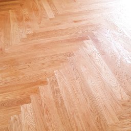 Ogólno-Budowlana - Ułożony parkiet w jodełkę francuską, jasne drewno, widoczne gniazdko elektryczne na szarej ścianie, świeżo pomalowane listwy przypodłogowe.